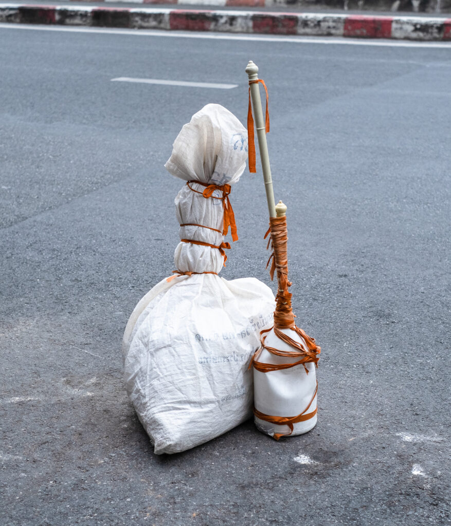 Photograph of two objects wrapped in orange ribbon on a road with red and white striped pavement visible across the other side. The object on the left is something in a white sack and the object on the right is a white plastic bottle with curtain poles coming out of the top.