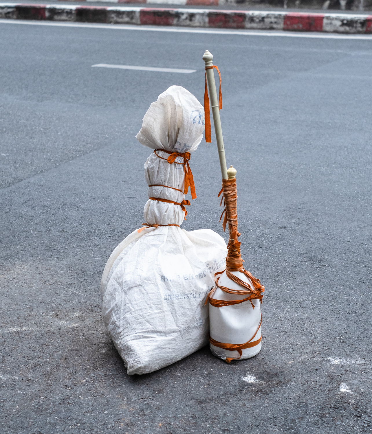 Photograph of two objects wrapped in orange ribbon on a road with red and white striped pavement visible across the other side. The object on the left is something in a white sack and the object on the right is a white plastic bottle with curtain poles coming out of the top.