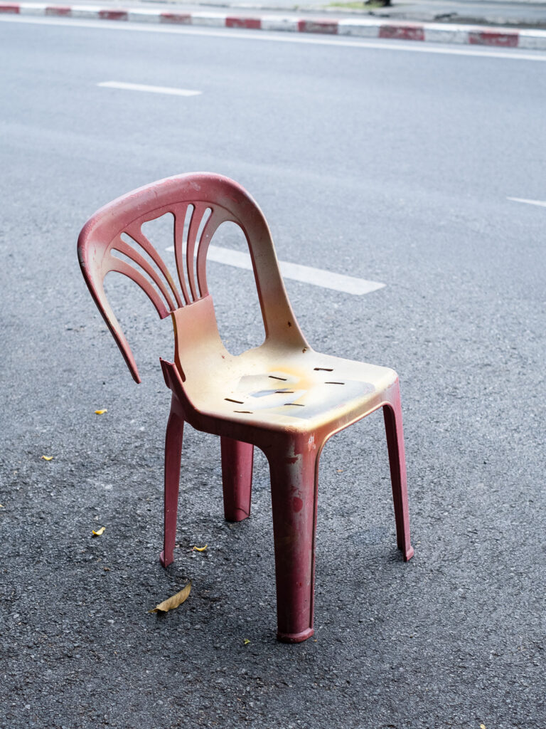 Photograph of a broken red plastic chair that has been sprayed with gold paint, in a road with white street markings behind and red and white painted curb in the distance.