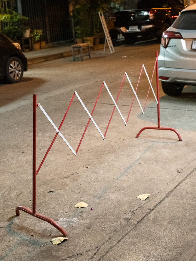 Photograph of an expandable metal barrier in red and white, blocking a space at the side of a road at night, with cars visible in the background.