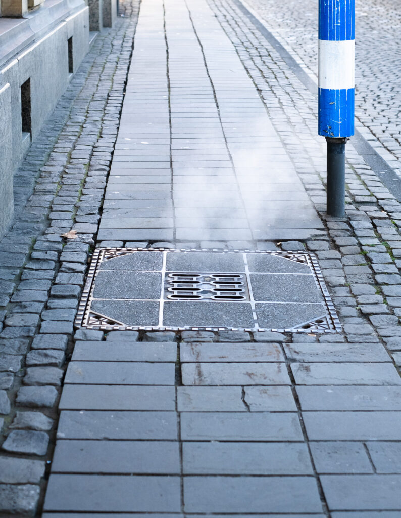 Photograph of a pavement with a utility cover with steam emerging from it and a pole with blue and white stripes on it. Cologne, Germany.