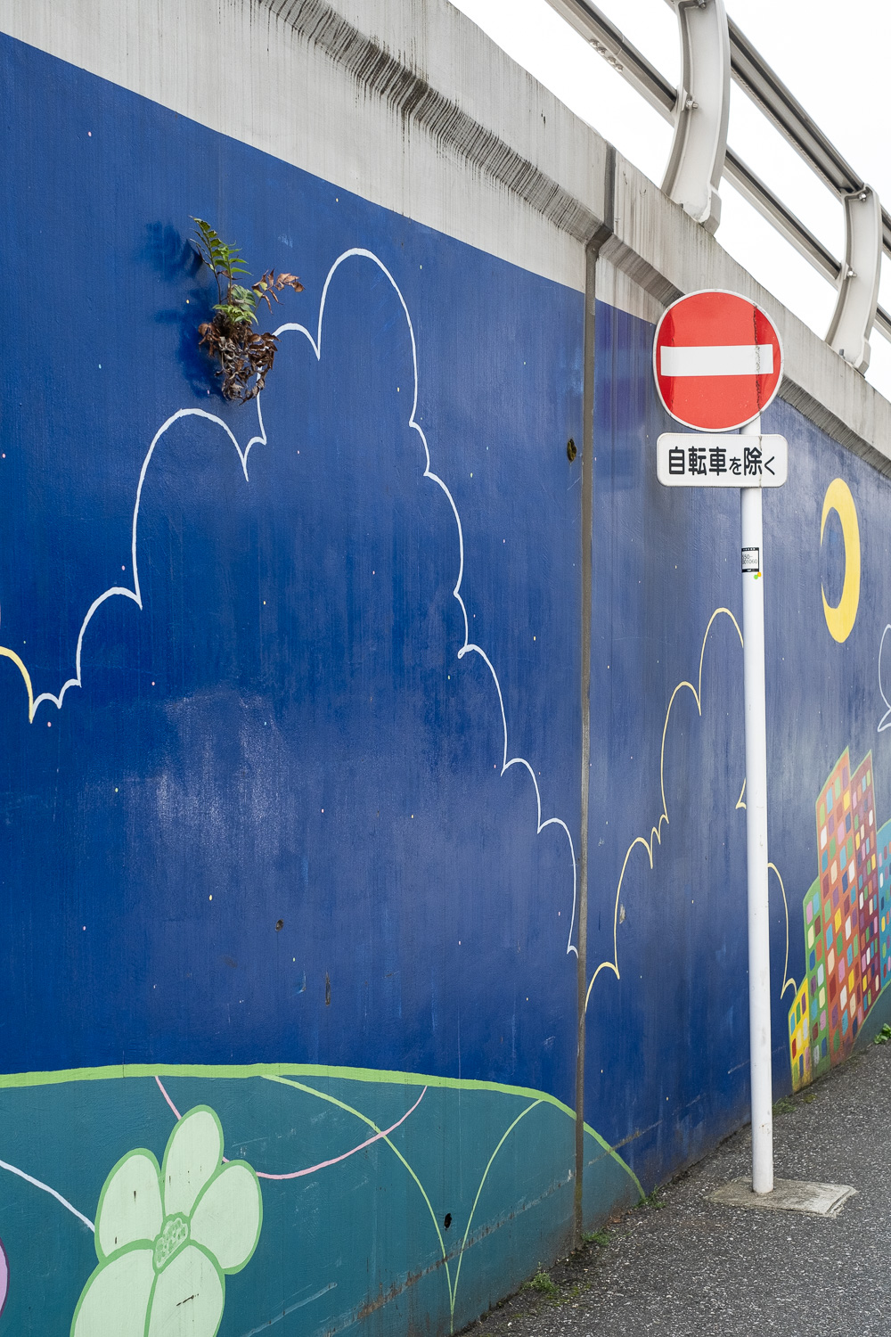 Photograph of a plant growing out of a wall that has been painted blue with a cloud outline, green and plants at the bottom and a moon in the distance to the right. A barrier is visible at the top of the frame and a stop sign is to the right.