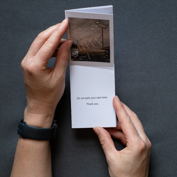 Photograph of 'Do not park your cars here', a zine by Jayne Lloyd, showing the A7 sized zine being held in front of a dark grey fabric background, it is being opened and 'Do not park your cars here. - Thank you.' is visible with a parking barrier at night visible upside down on the page that's being folded back.