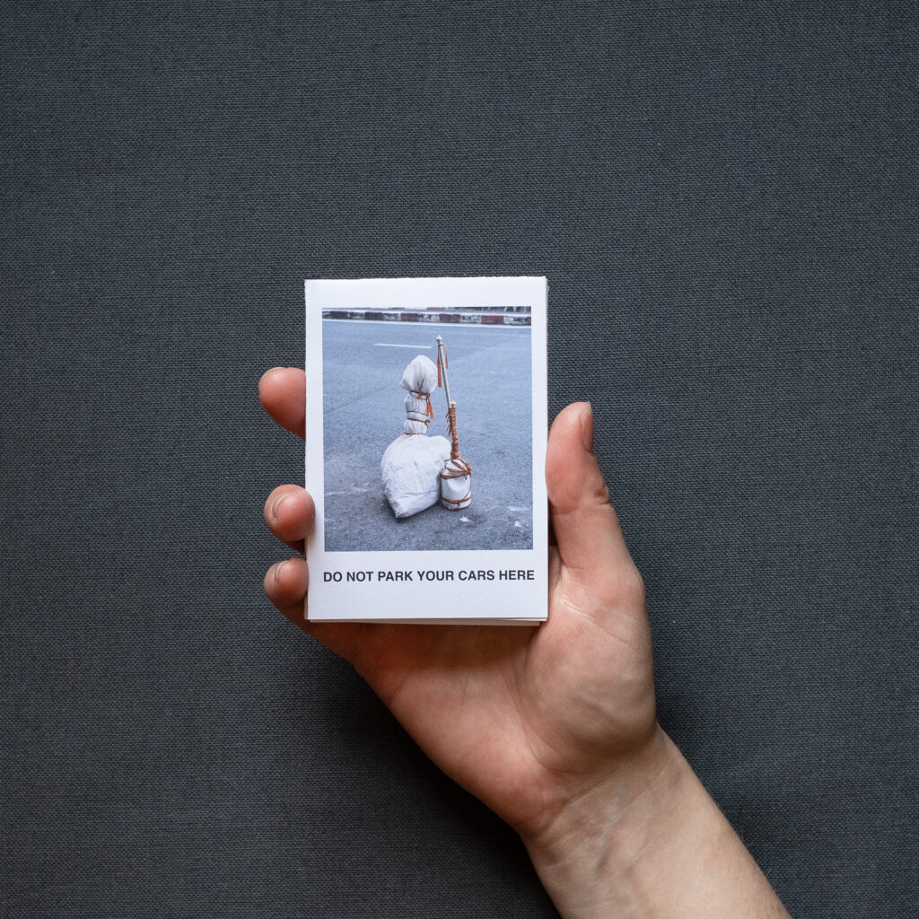 Photograph of 'Do not park your cars here', a zine by Jayne Lloyd, showing the A7 sized zine being held in front of a dark grey fabric background.