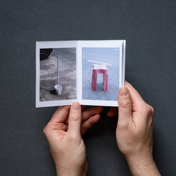 Photograph of 'Do not park your cars here', a zine by Jayne Lloyd, showing the A7 sized zine being held in front of a dark grey fabric background. The page on the left shows a white bucket with a light blue pole sticking out of it and the page on the right has a pink plastic stool.