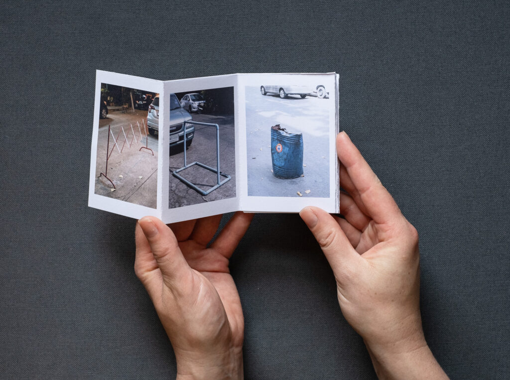 Photograph of 'Do not park your cars here', a zine by Jayne Lloyd, showing the A7 sized zine being held in front of a dark grey fabric background.