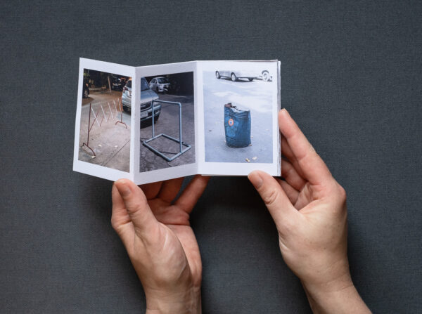 Photograph of 'Do not park your cars here', a zine by Jayne Lloyd, showing the A7 sized zine being held in front of a dark grey fabric background. On the far left is a photograph of a parking barrier at night, in the centre is a photograph of a barrier made of light blue plastic pipes and on the right is a photograph of a battered metal blue barrel in the road.