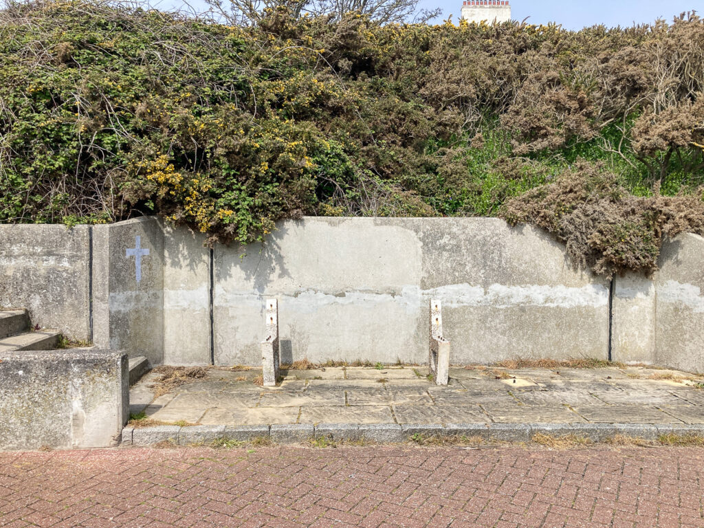 Photograph of a paved area with the legs of a concrete bench, with the seat area missing. Behind are plants on a hill.