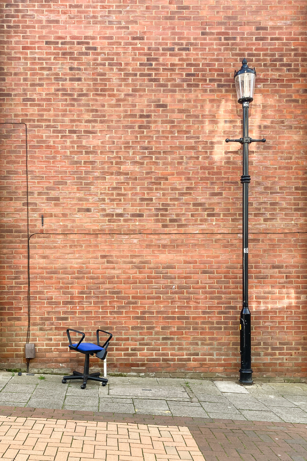 Photograph of a brick wall with a black old fashioned lampost to the right and the lower part of an office chair with a blue seat to the left, on a sloped paved road.