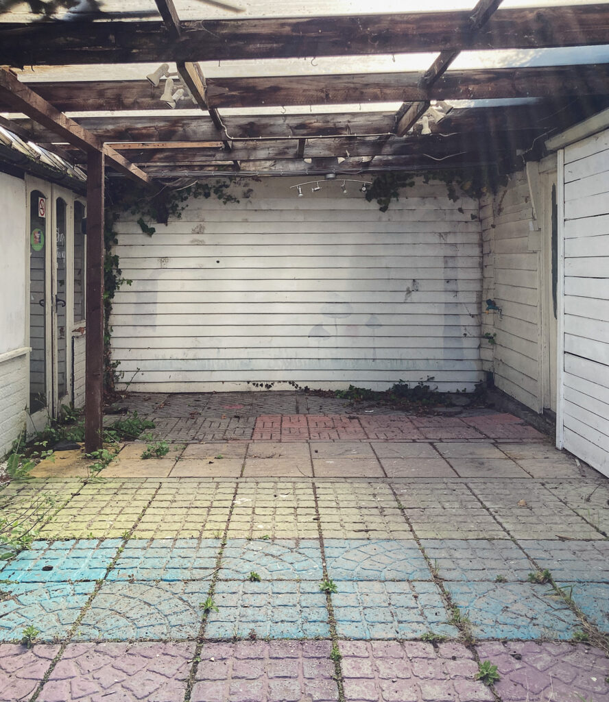 Photograph of a covered outdoor space with a wooden wall at the far end, with a scene of trees, grass and mushrooms painted over in white and rainbow coloured paving in the foreground.