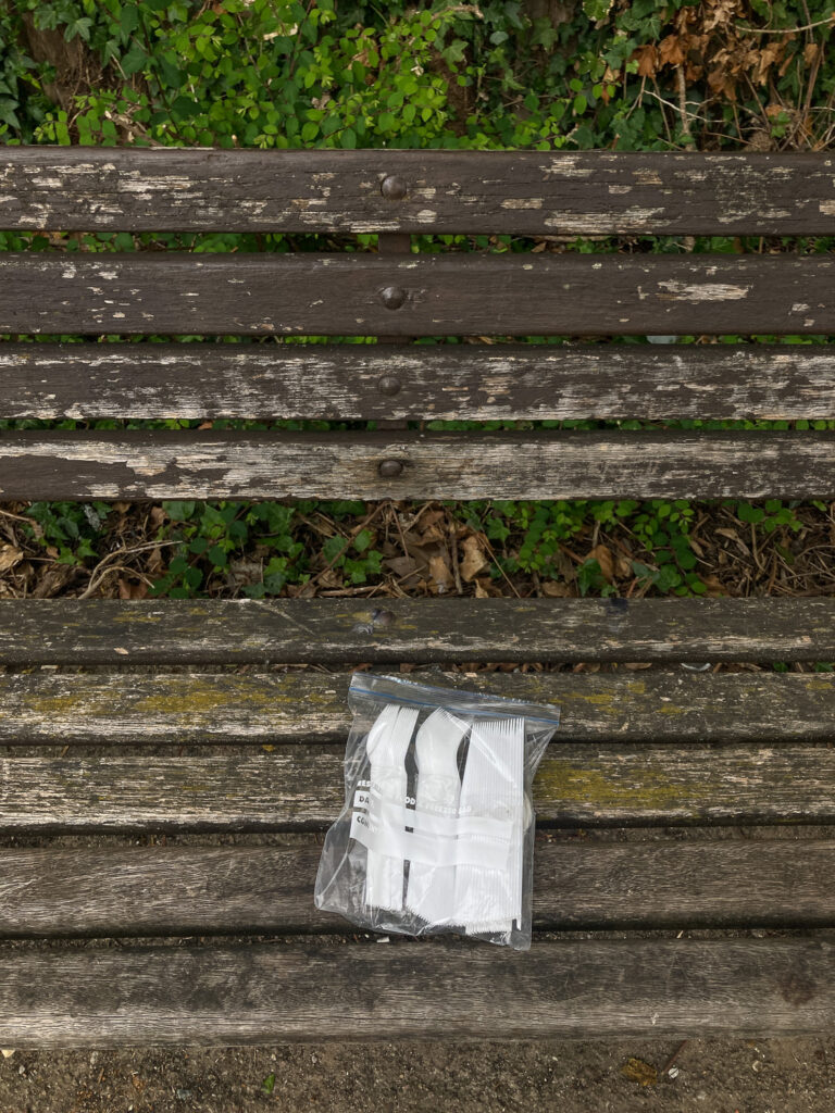 Photograph of a sandwich bag filled with plastic cutlery on a worn wooden bench, with plants behind.