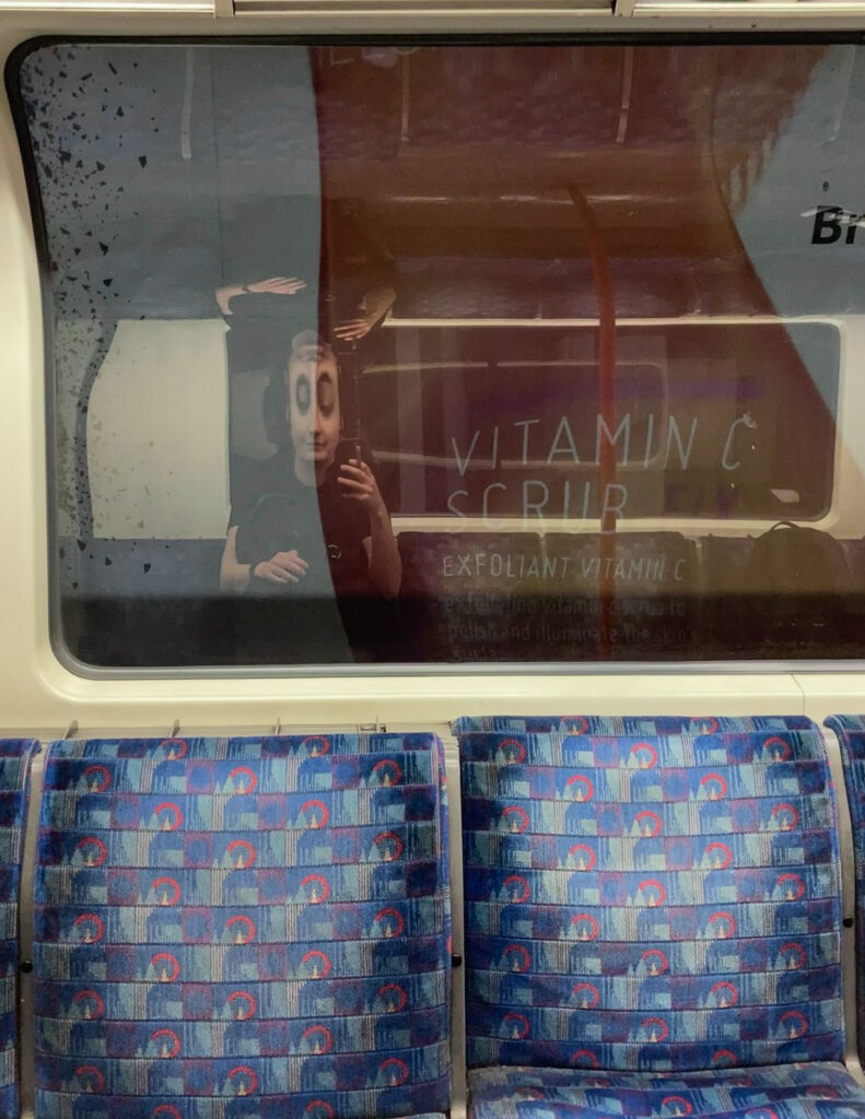 Photograph of a window and seats in a tube carriage, with a reflection of a white woman taking a selfie, her reflection distorted so her eyes look big and black as she's also reflected upside down above due to the curved surface. An advert through the window says 'VITAMIN C SCRUB, EXFOLIANT VITAMIN C'