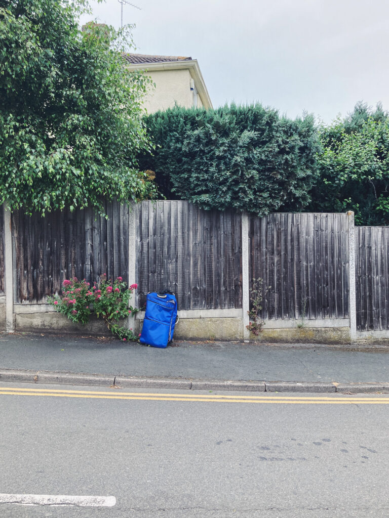 Photograph of a street with road in the foreground, a blue luggage bag on a pavement leaning against a wooden fence with buses behind.