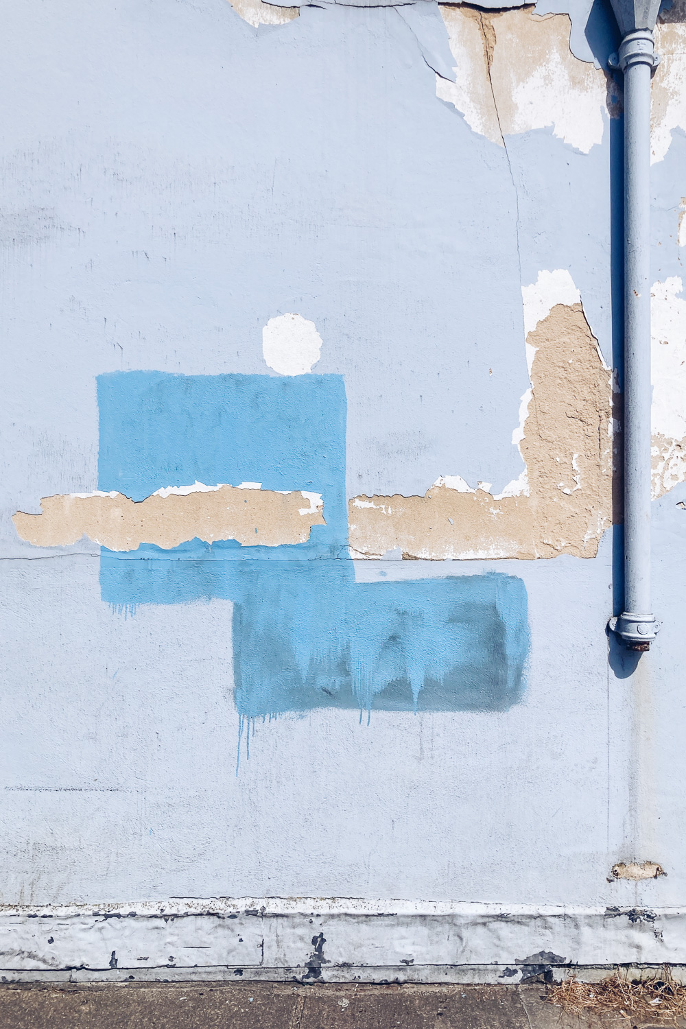 Photograph of a light blue wall with patches of another blue and peeled of plaster in the centre and a broken drainpipe to the right.