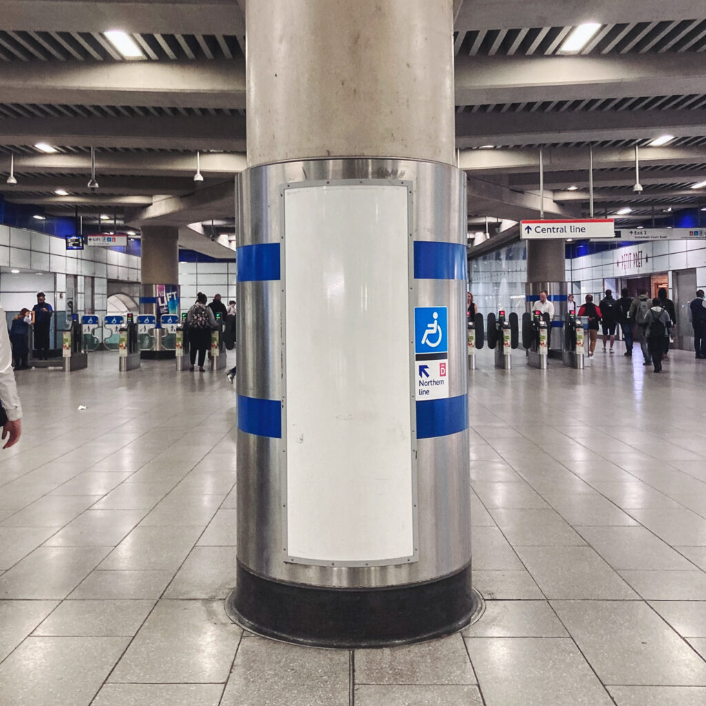 Photograph of an empty notice area on a pillar at Tottenham Court Road station, with people walking in the background.