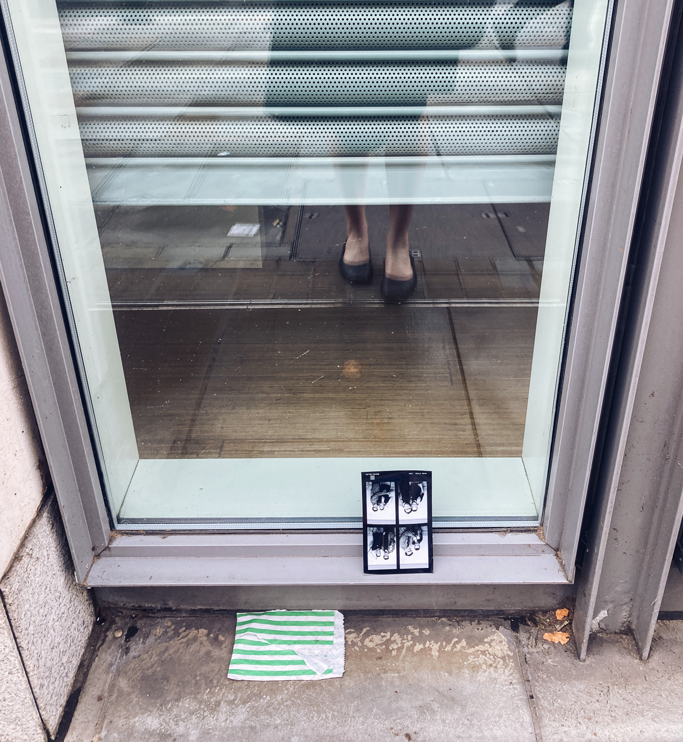 Photograph  floor-length window with a reflection of a white woman's legs and shoes, and an upside down Photo Booth style print with 4 images of 2 people posing for pictures