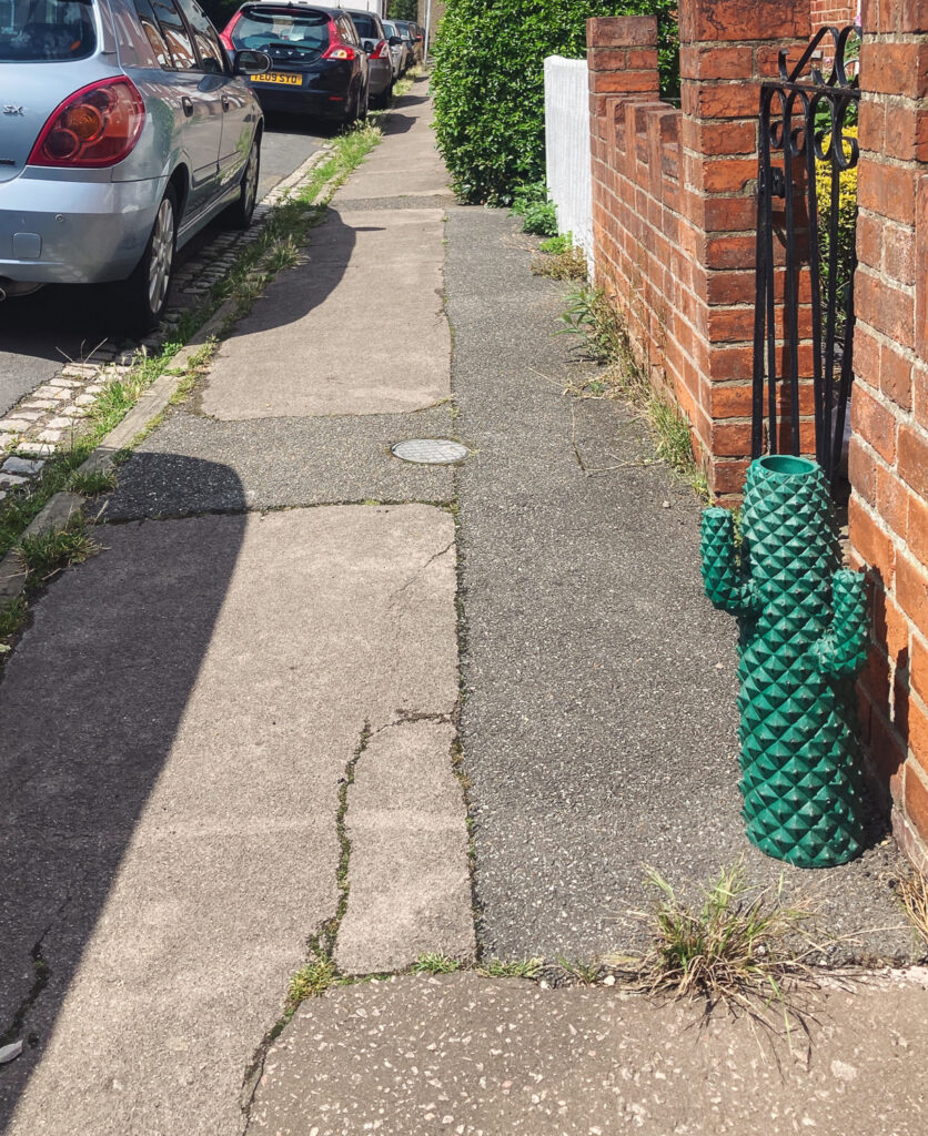 Photograph of a pavement with a planter in the shape of a cactus to the right, in front of a wall and cars parked along the road to the left and into the distance.