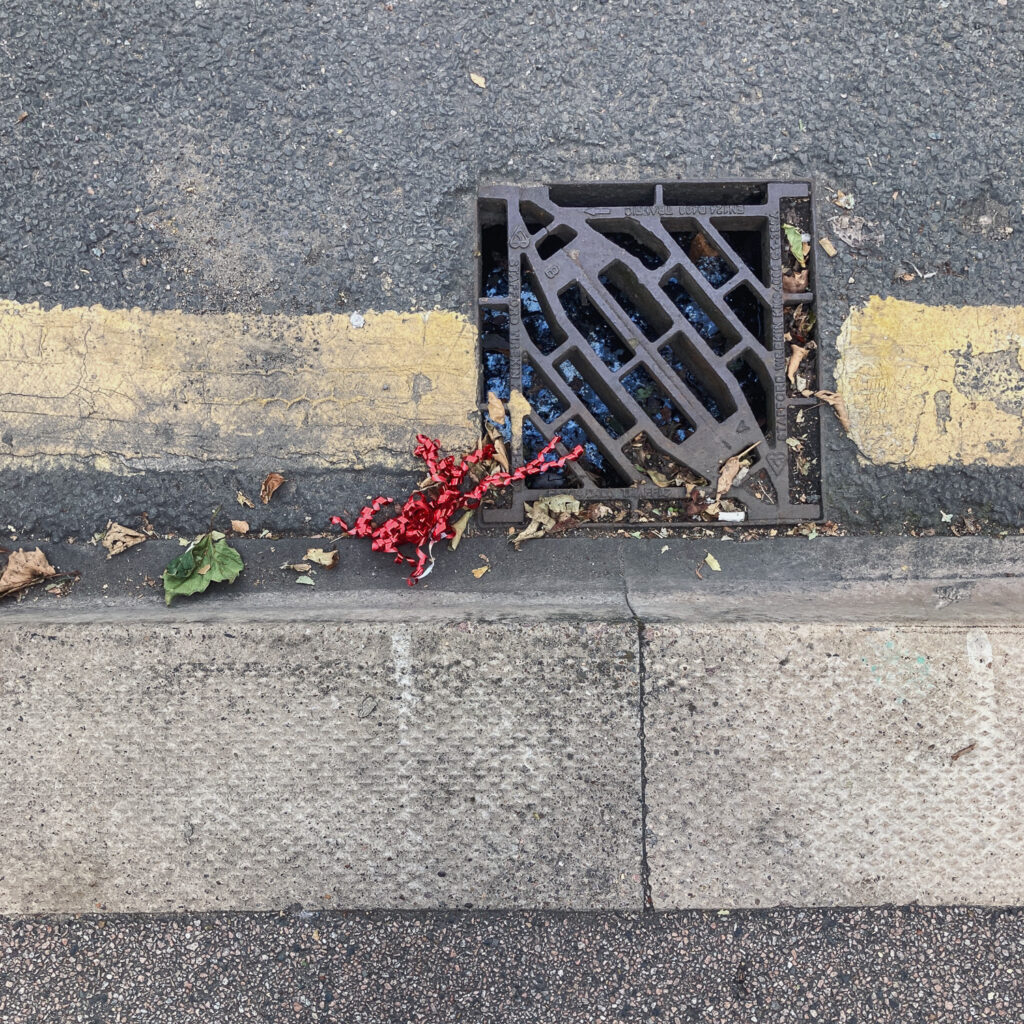 Photograph of red curly ribbon in a gutter at the side of the road, with pavement in the lower portion, a drain and yellow painted line towards the centre.