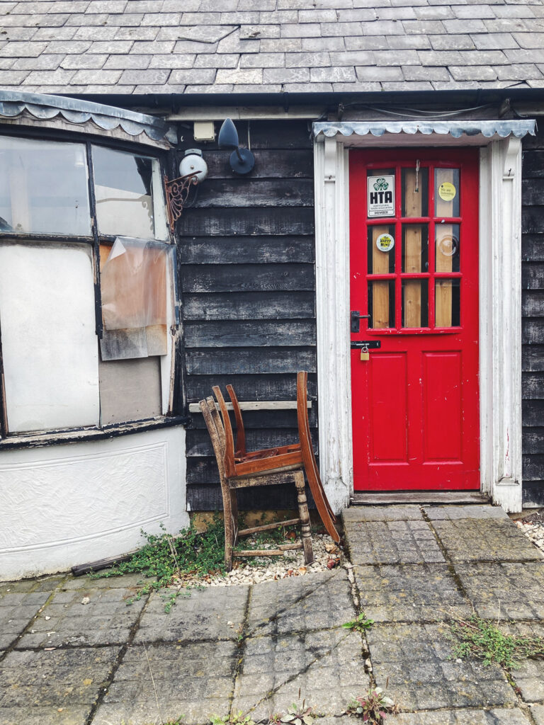 Photograph of two chairs, one upside down on top of the other, in front of a building with red door to the left, boarded up bag window to the left and paving slabs in the foreground.