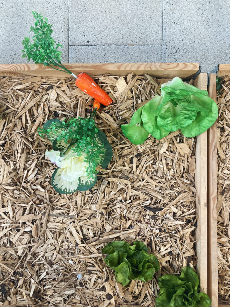 Photograph looking down at a wooden box filled with wooden chips, with plastic lettuces and carrots on top, with paving slabs visible at the top portion of the frame.