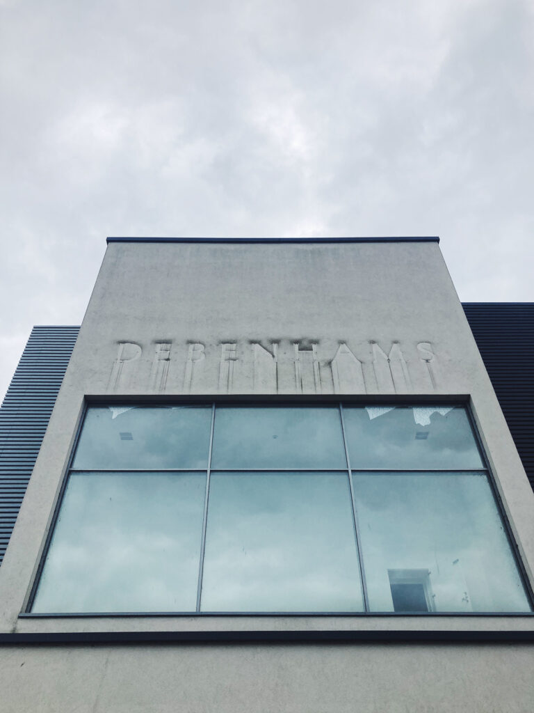 Photograph looking up at a former Debenhams building, a concrete building with the word 'DEBENHAMS' visible as dirt from around the signage that was there before, with a large window below, showing an open doorway in the right-hand pane, with a cloudy sky above.