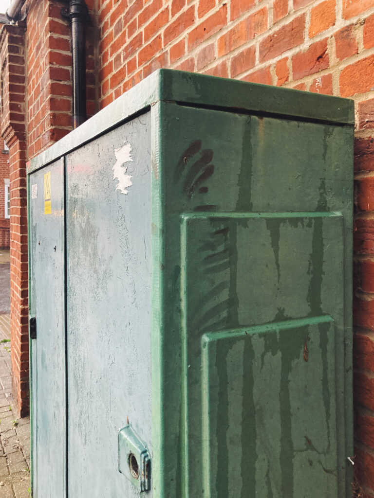 Photograph of a green telephone connection box in front of a brick walled-building, with greasy hand smears on the side.