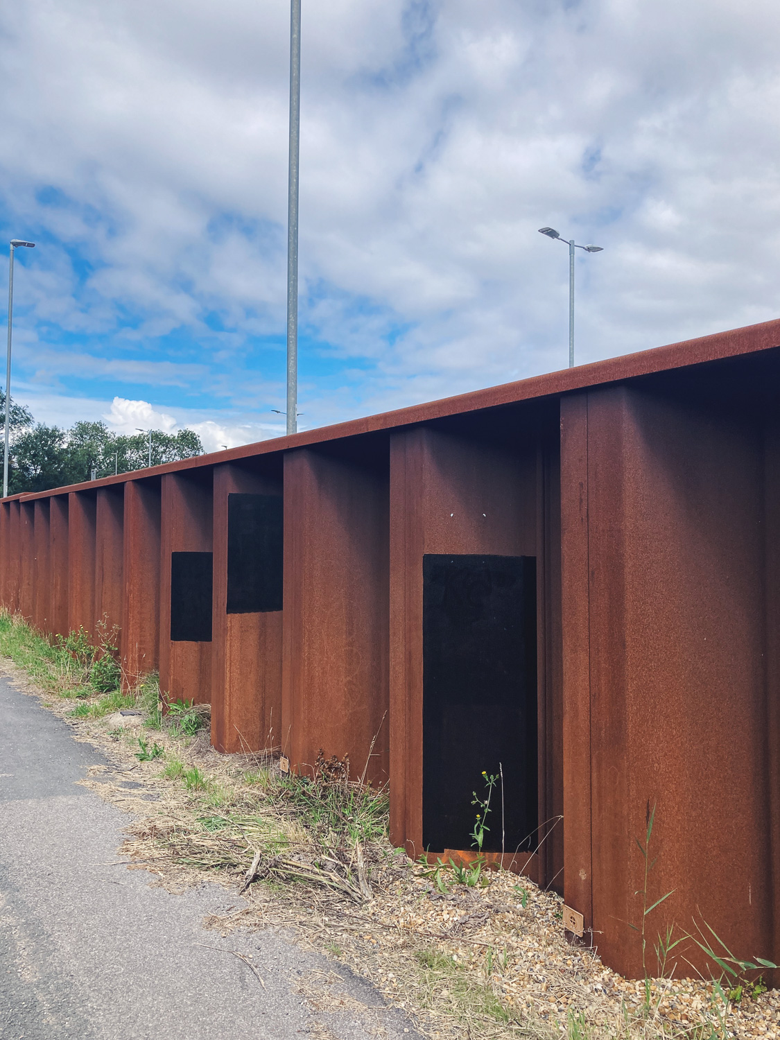 Photograph of black rectangles painted onto rusty metl fencing, with road to the lower left and blue sky and clouds above, with lampposts in the distance.