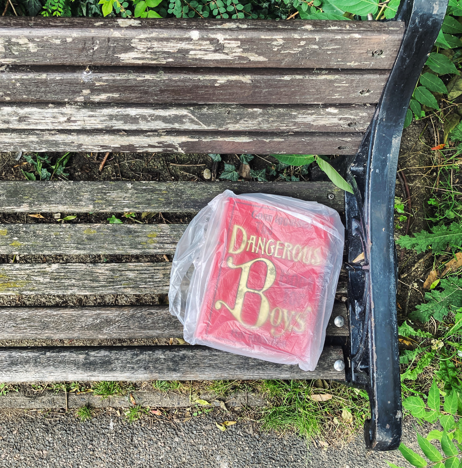 Photograph looking down at a worn wooden bench with metal arm rest, upon which is a plastic bag with a book in it, and ‘Dangerous Boys’ in yellow text on a red background visible through the plastic.
