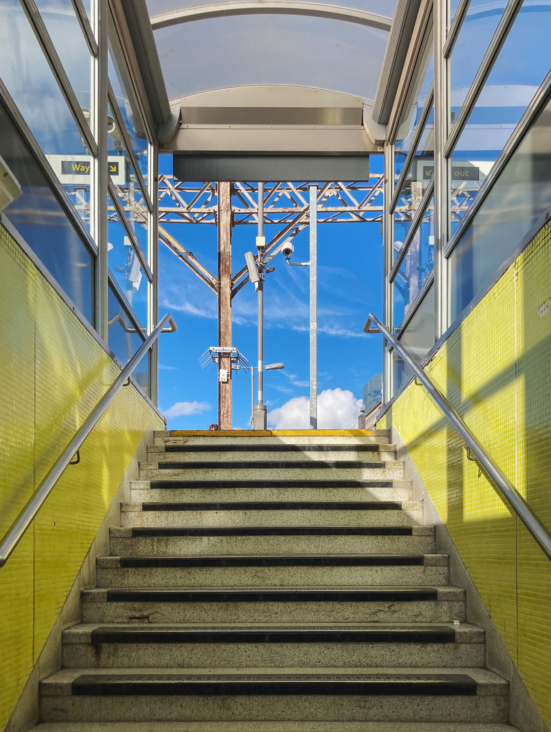 Photograph looking up a staircase with yellow tiled walls with metal handrails either side and glass top and side above, with metal structures and blue sky in the distance.
