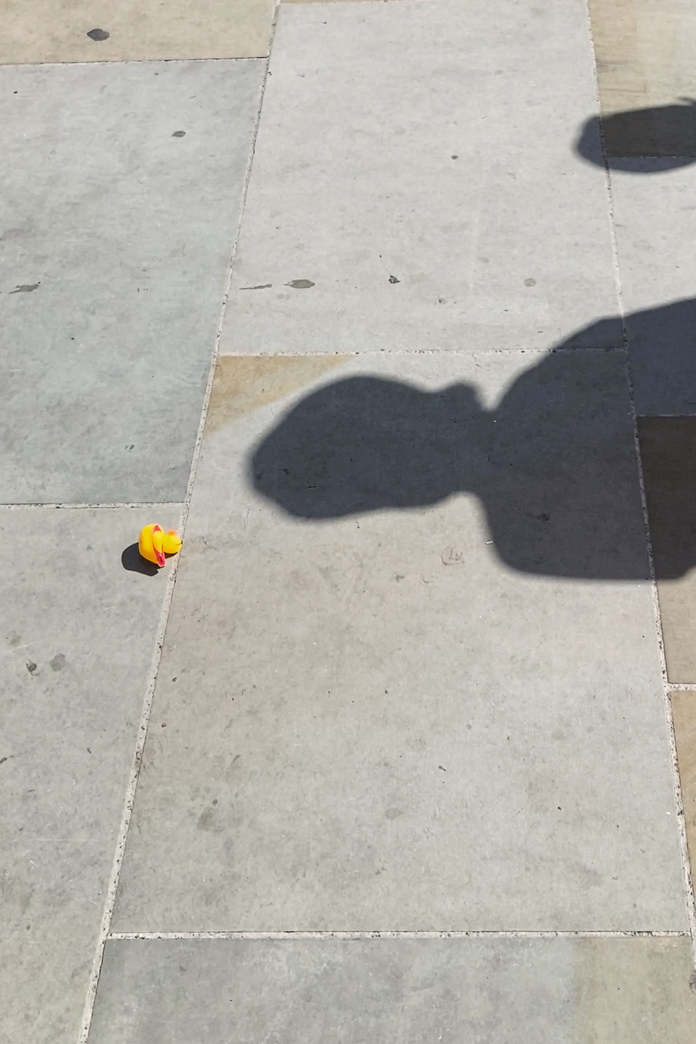 Photograph of a tiled pavement with a yellow rubber duck to the left and shadows of people walking by in the centre of the image.