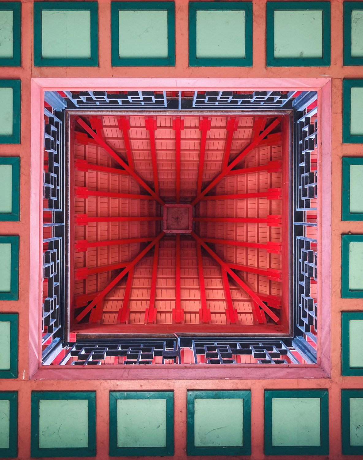 Photograph looking up at the ceiling in the Chinese Pagoda in Victoria Park, with green squares separated by pink lines around the edge, a geometric pattern metal barrier within that and red and pink lines of the roof within that.