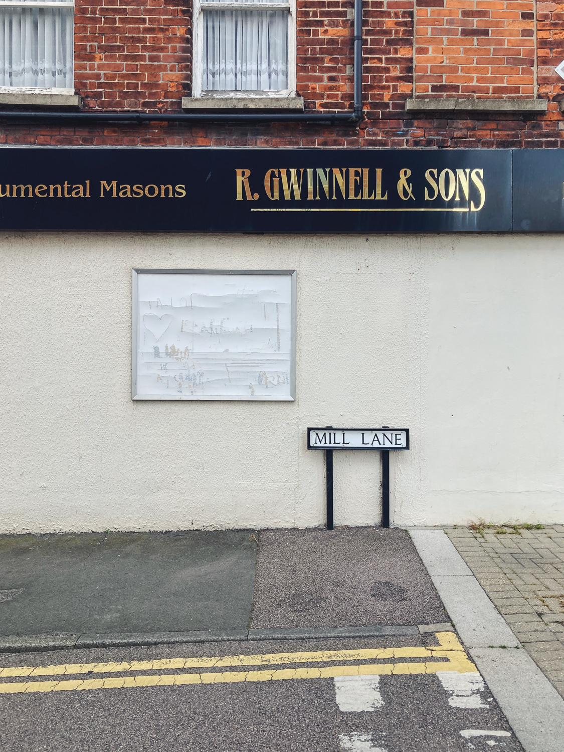 Photograph of an empty noticeboard on a wall with a street name 'MILL LANE' to the right, '...umental Masons, R. GWINNELL & SONS' on a sign above and windows and brick wall above that, with pavement and double yellow lines at the bottom of the frame.