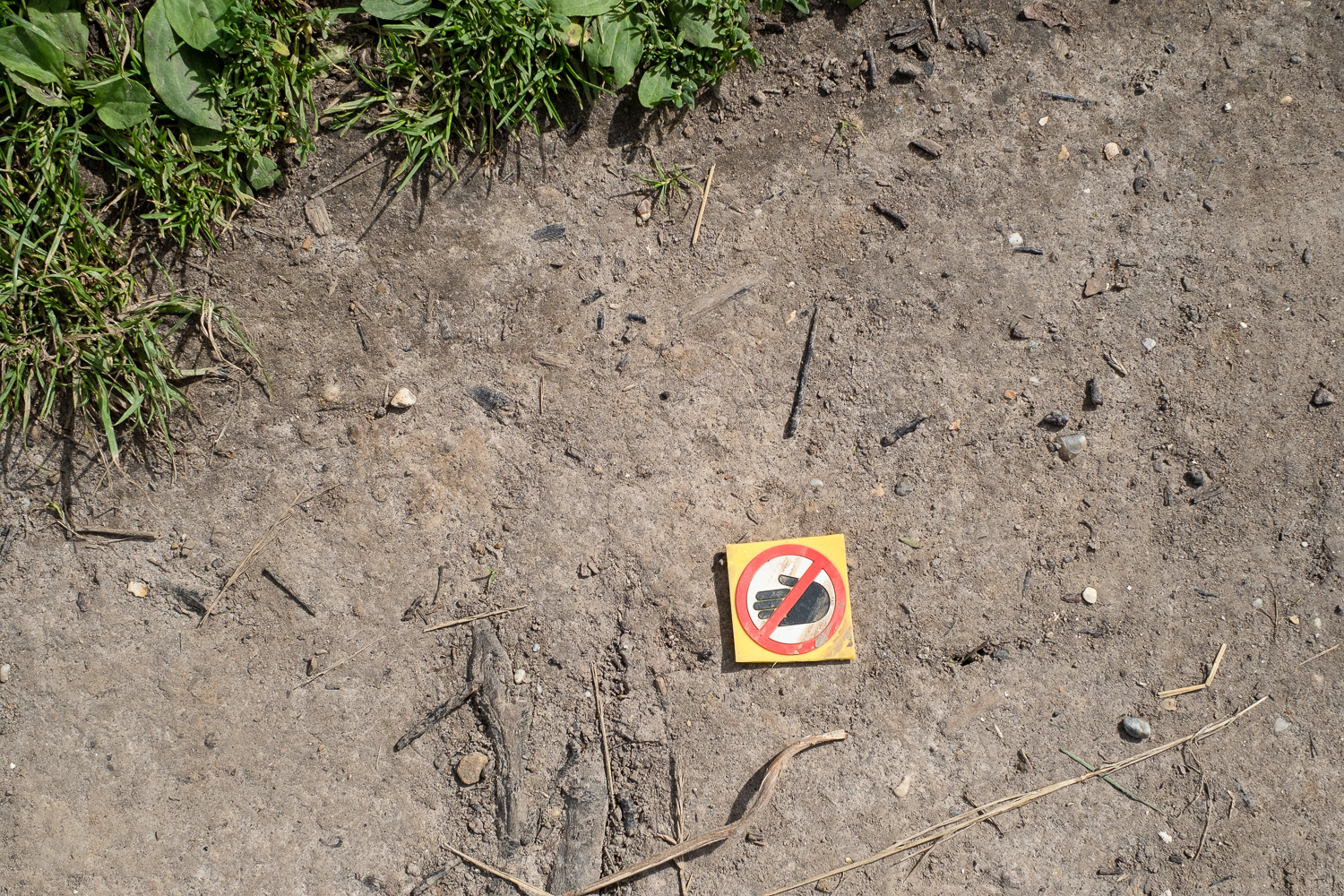 Photograph of a plastic sign showing a black hand shape on a white background, within a red circle with a red line across it, with a yellow border around it. All on a dusty path with grass to the top left of the frame. Walton-on-the-Naze, Essex