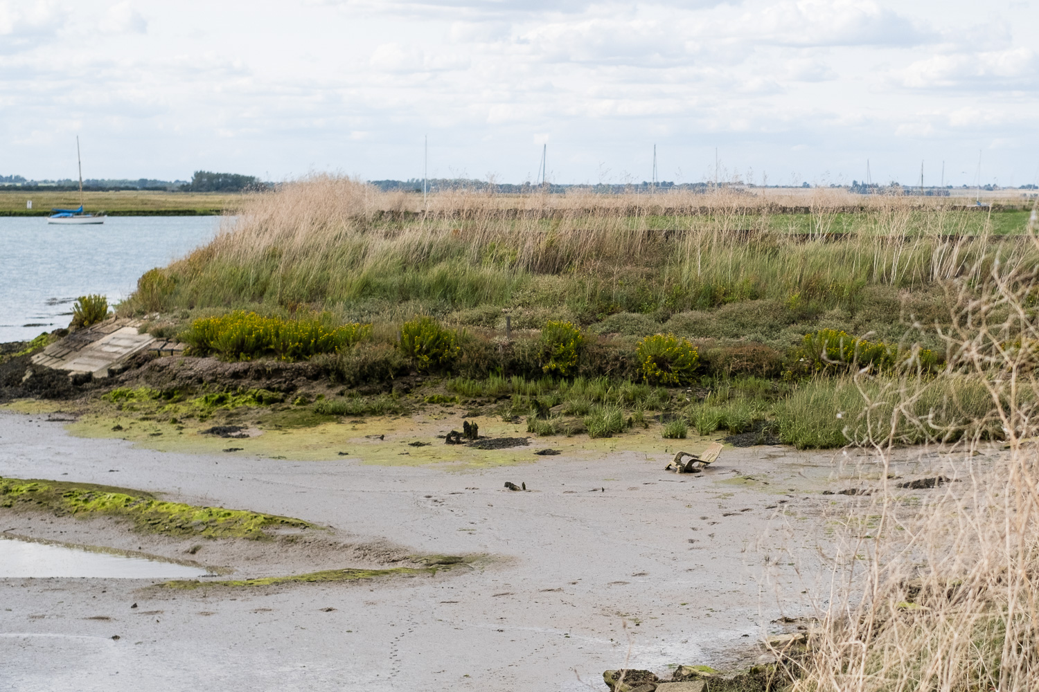 Photograph of a plastic garden chair embedded in mud with footprints to it, surrounded by grass  and sailing yacht visible in the background. Walton-on-the-Naze, Essex