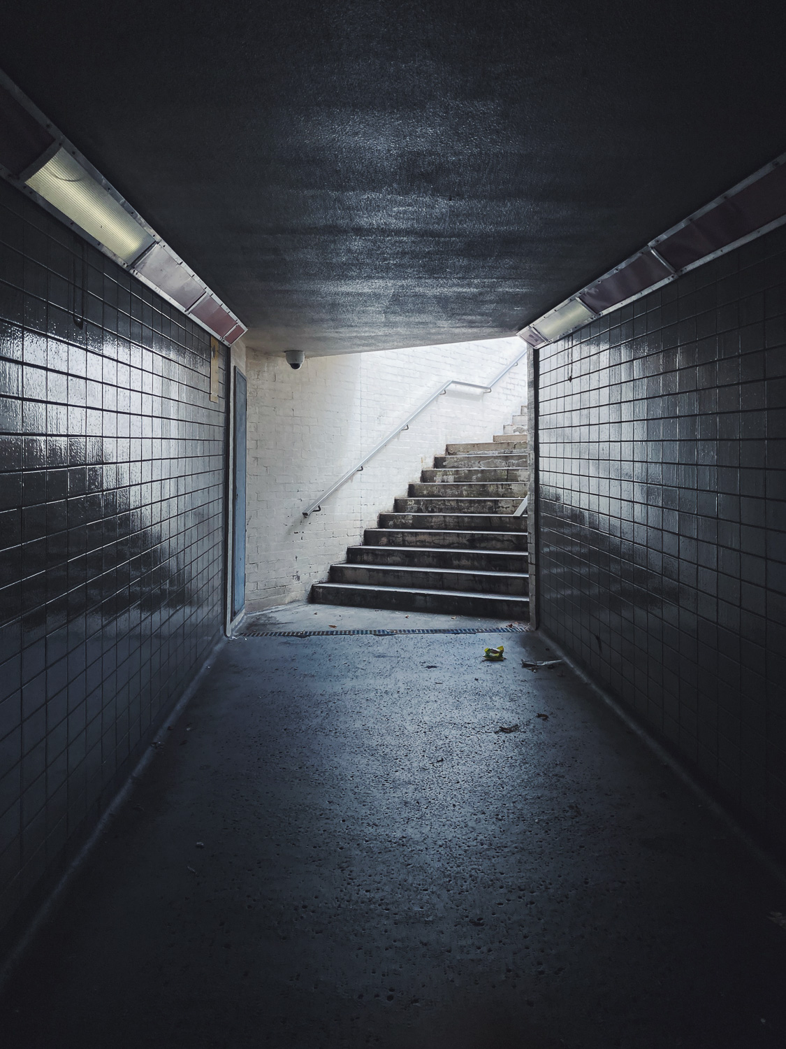 Photograph in an underpass with steps leading up to the right in the centre, tiled walls either side and lights along the top of each.