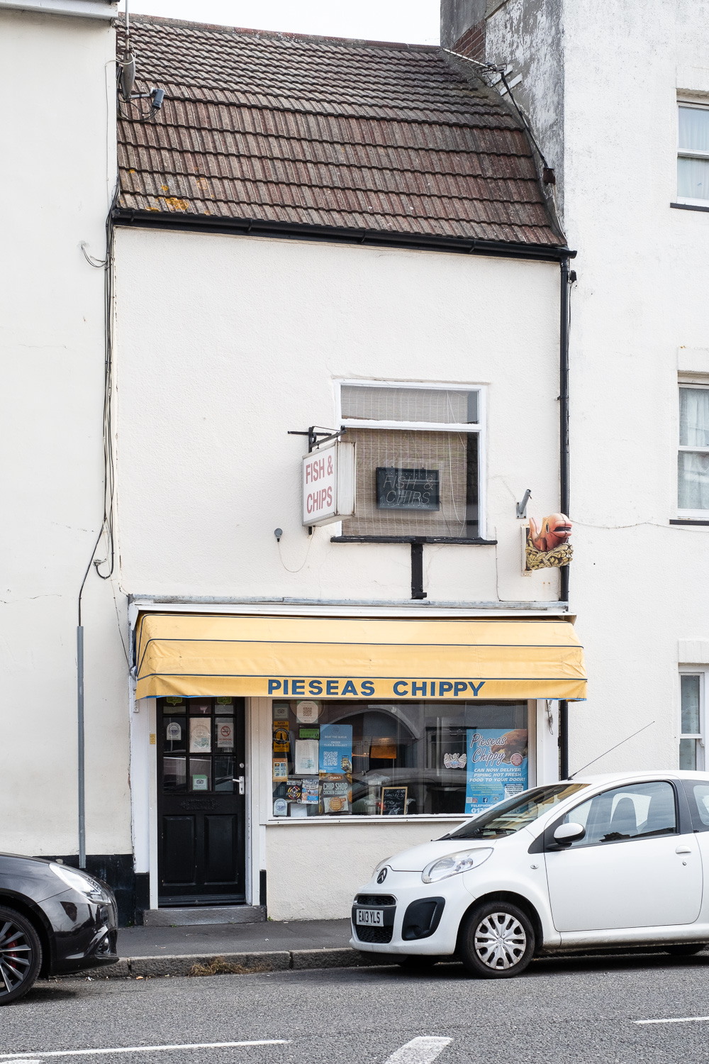 Photograph of Pieseas Chippy, Harwich, Essex, UK with a yellow canopy in front of the shop with door and a wide window beneath, cars in the road in front and window and 'Fish & Chips' sign above.