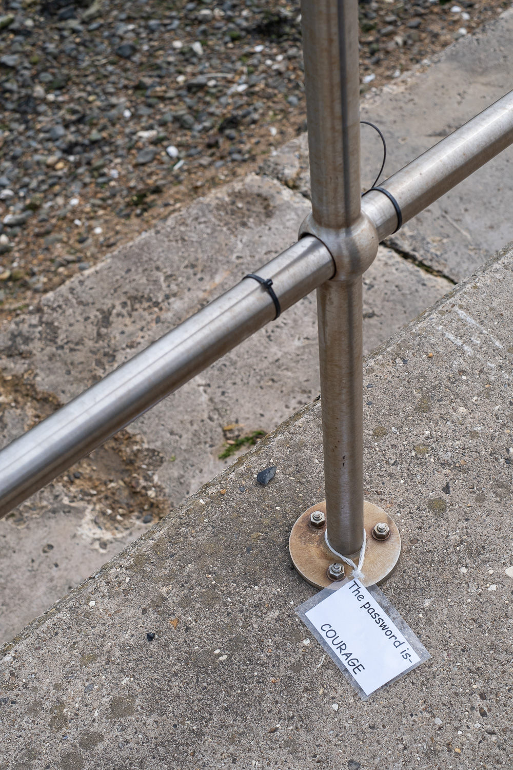 Photograph of a laminated sign saying 'The password is- COURAGE', on a concrete floor, attached to a metal railing. Harwich, Essex, UK