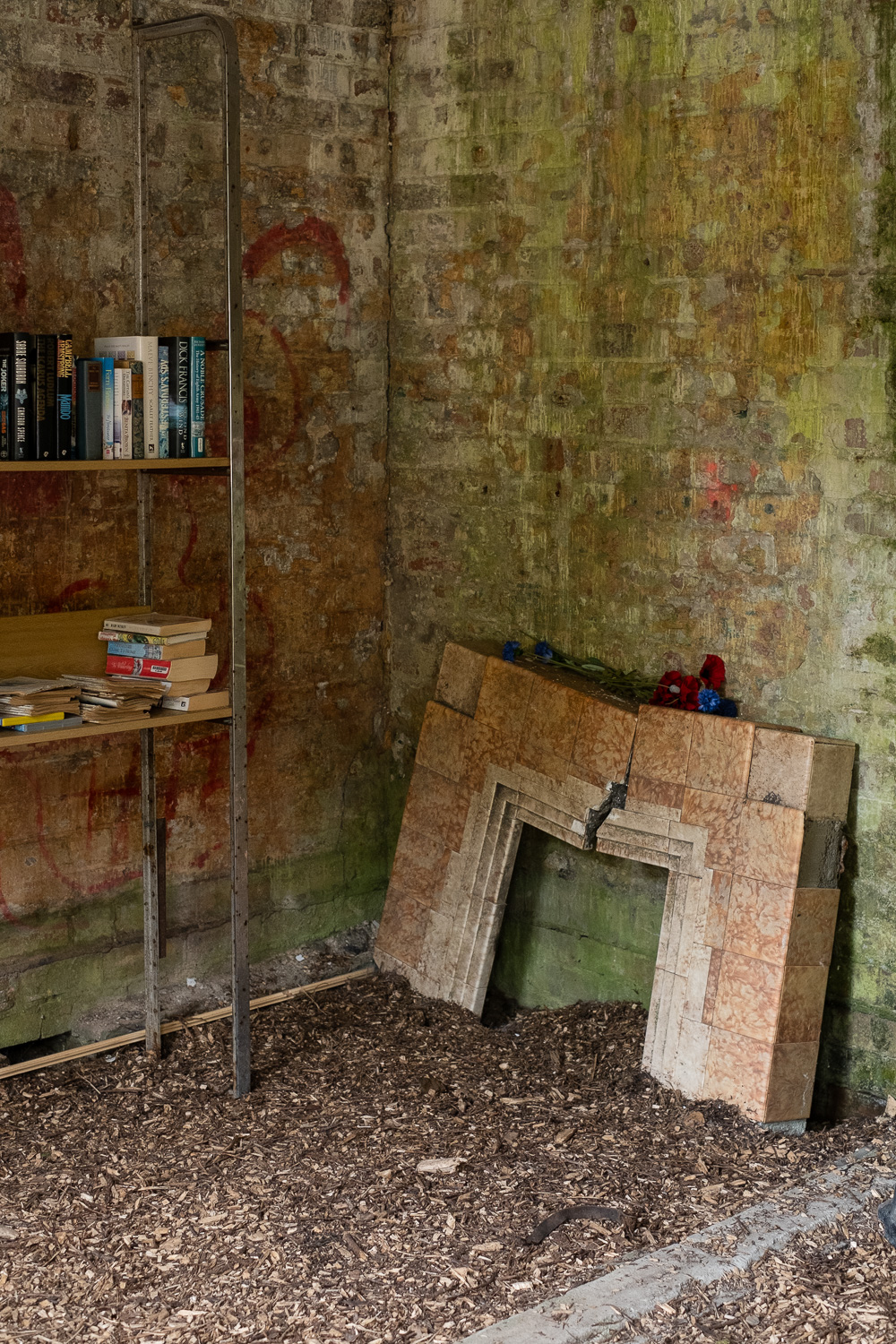 A broken tiled mantelpiece in a bare bricked wall, with bark on the floor and bookshelves with a few books to the left. Harwich, Essex, UK