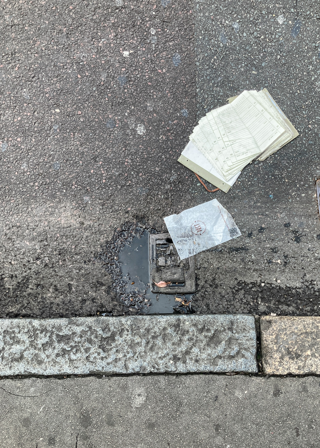 Photograph of a flattened diary in a road with a grate with a puddle and Pret paper bag in the centre, with curb and pavement visible in the lower portion of the frame.