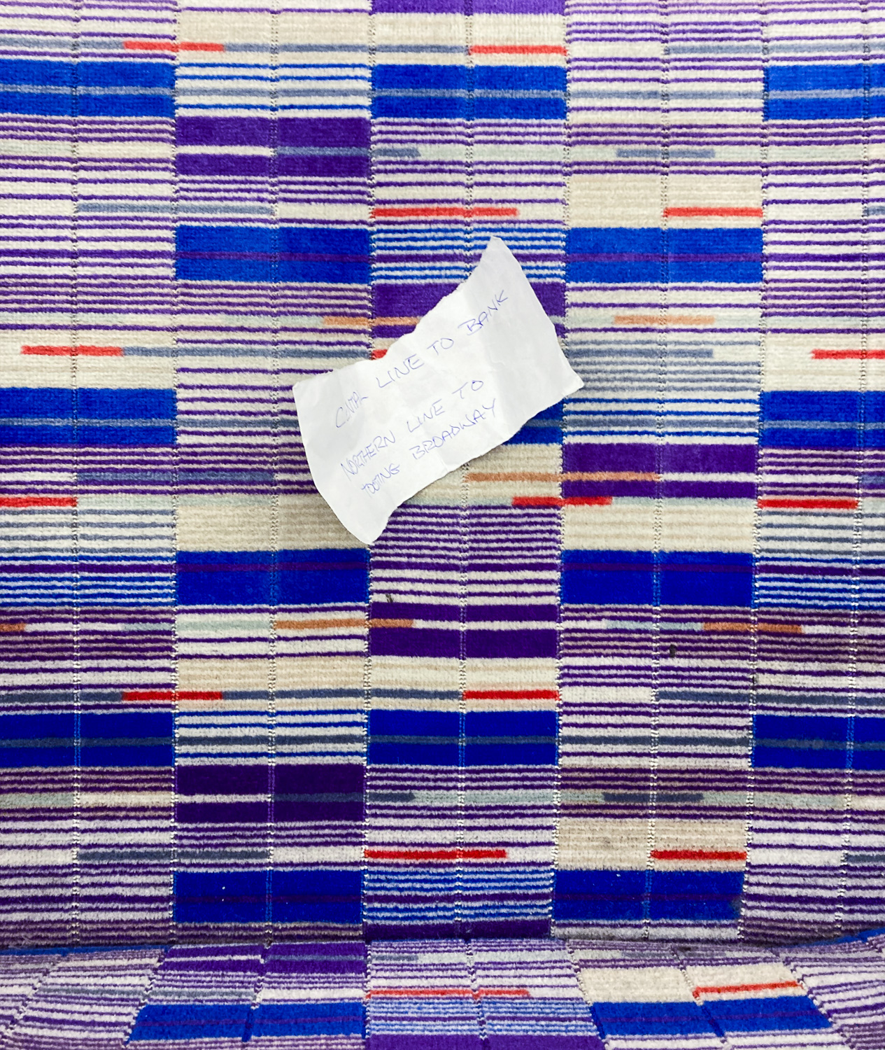 Photograph of a scrap of paper with 'CENTRAL LINE TO BANK, NORTHER LINE TO EALING BROADWAY' on Elizabeth Line patterned seat fabric.