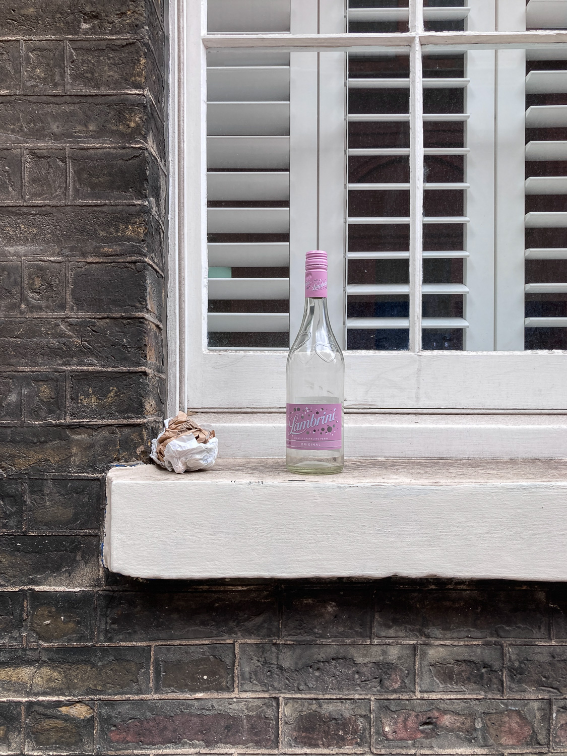Photograph of an empty bottle with a pink label saying 'Lambrini' on a window ledge, next to a screwed up paper bag next to it and a window with shutters behind, in a brick wall.