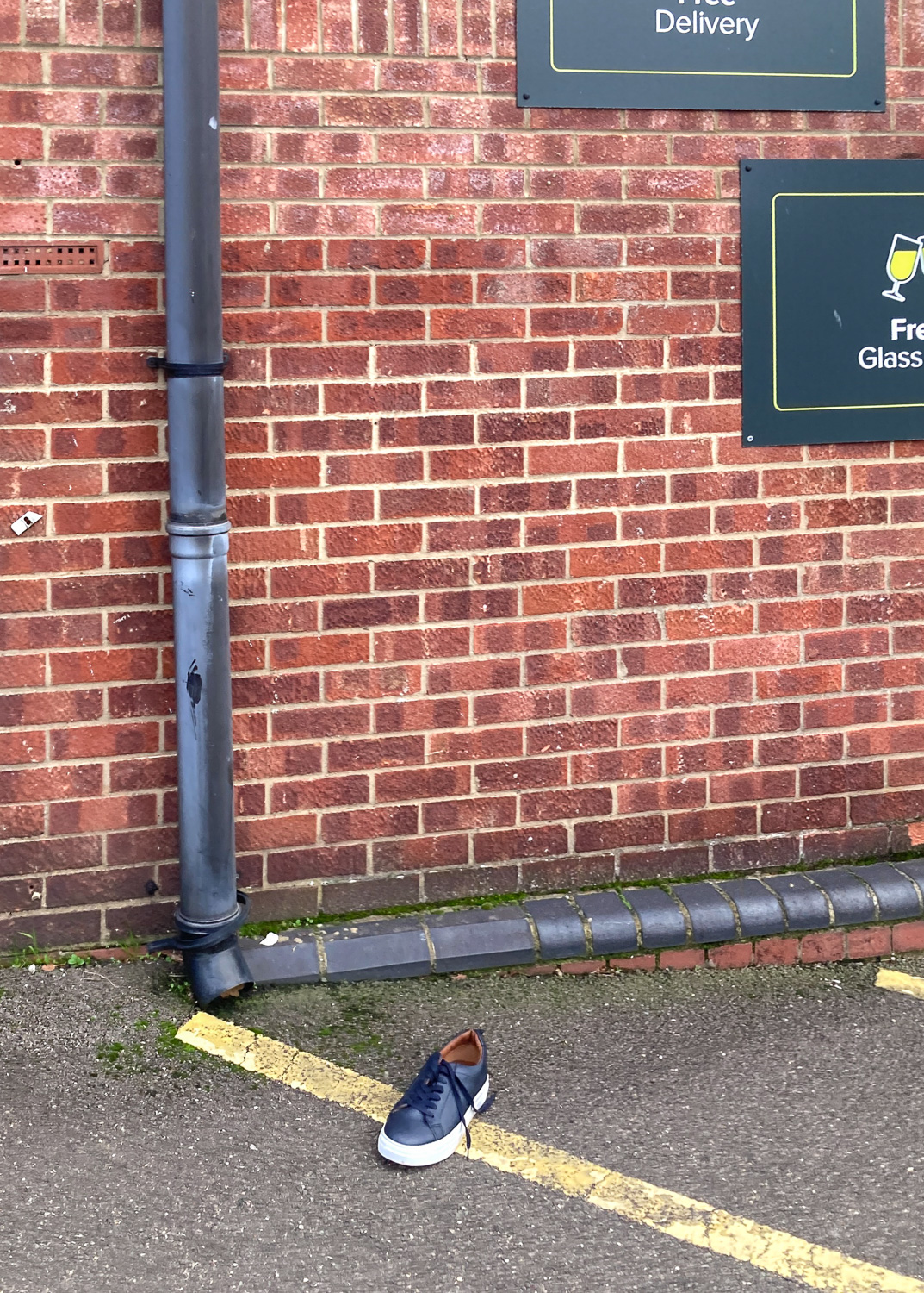 Photograph of a single shoe on the floor, on a diagonal yellow painted line, with a brick wall behind that has a drainpipe to the left and 2 partly visible signs to the top right.