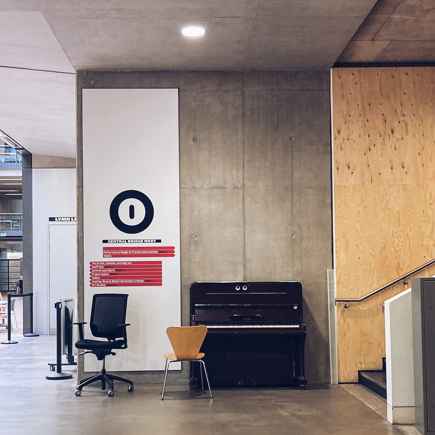 Photograph of an area at the bottom of som stairs, with a sign on the wall, a black office chair, a wooden chair and an upright piano with two comic eyes stuck on it.