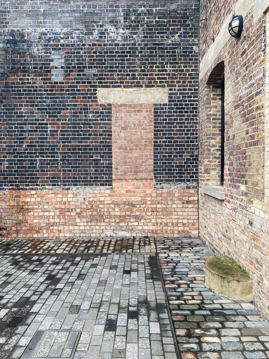 Photograph of a bricked-in window in a brick wall with contrasting coloured bricks, with brick ground in front and another brick wall with a recessed window and wall-mounted light to the right.