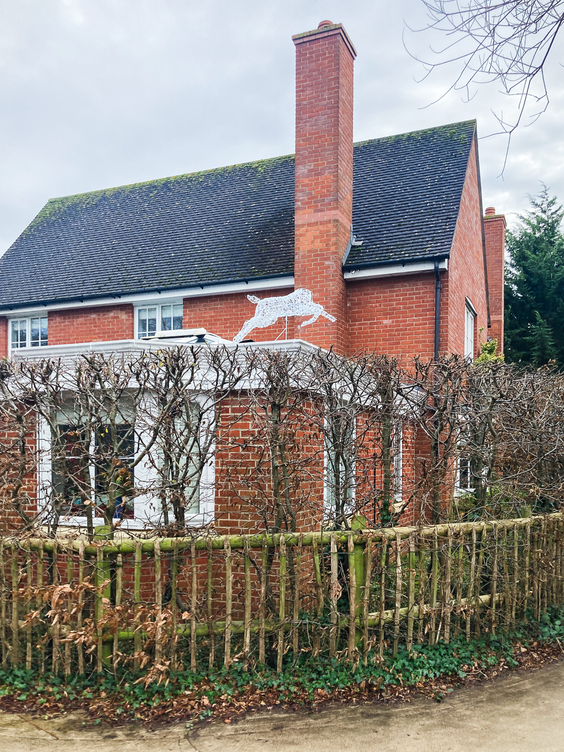 Photograph of a brick house with a tall chimney, surrounded by a wooden fence with trimmed trees. Above the trees, mounted on the low roof of a part of the house is a large deer-shaped Christmas light decoration, with its head missing.