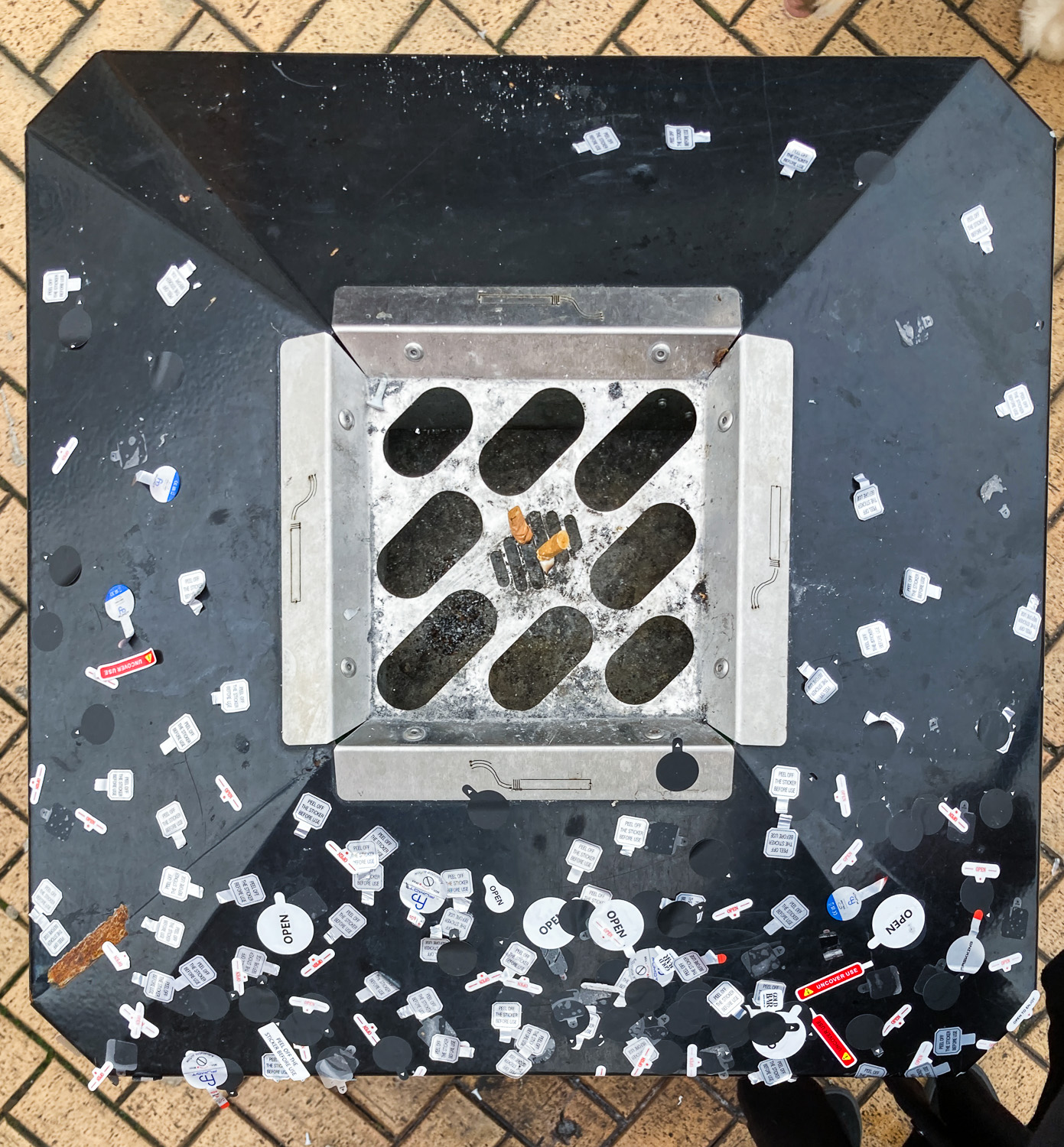 Photograph looking down on a public waste bin, the edges of which are covered in stickers from Vape packaging. In the centre is a metal grid for disposing of cigarettes, and the brick pavement can be seen below, around the edges of the frame.