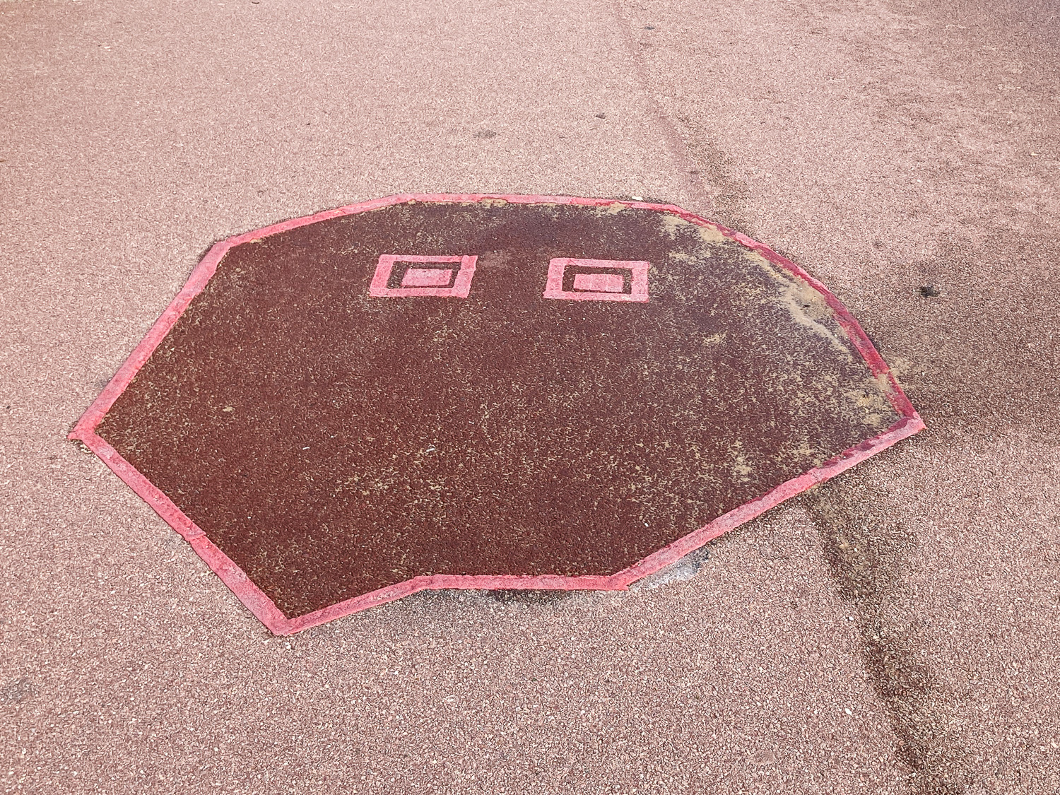 Photograph of a red tarmac pavement that has been patched up with a darker red colour and bright red tape-like areas that resemeble the shape of a ghost with square eyes. Sand is visible across the surface as it is by the beach.