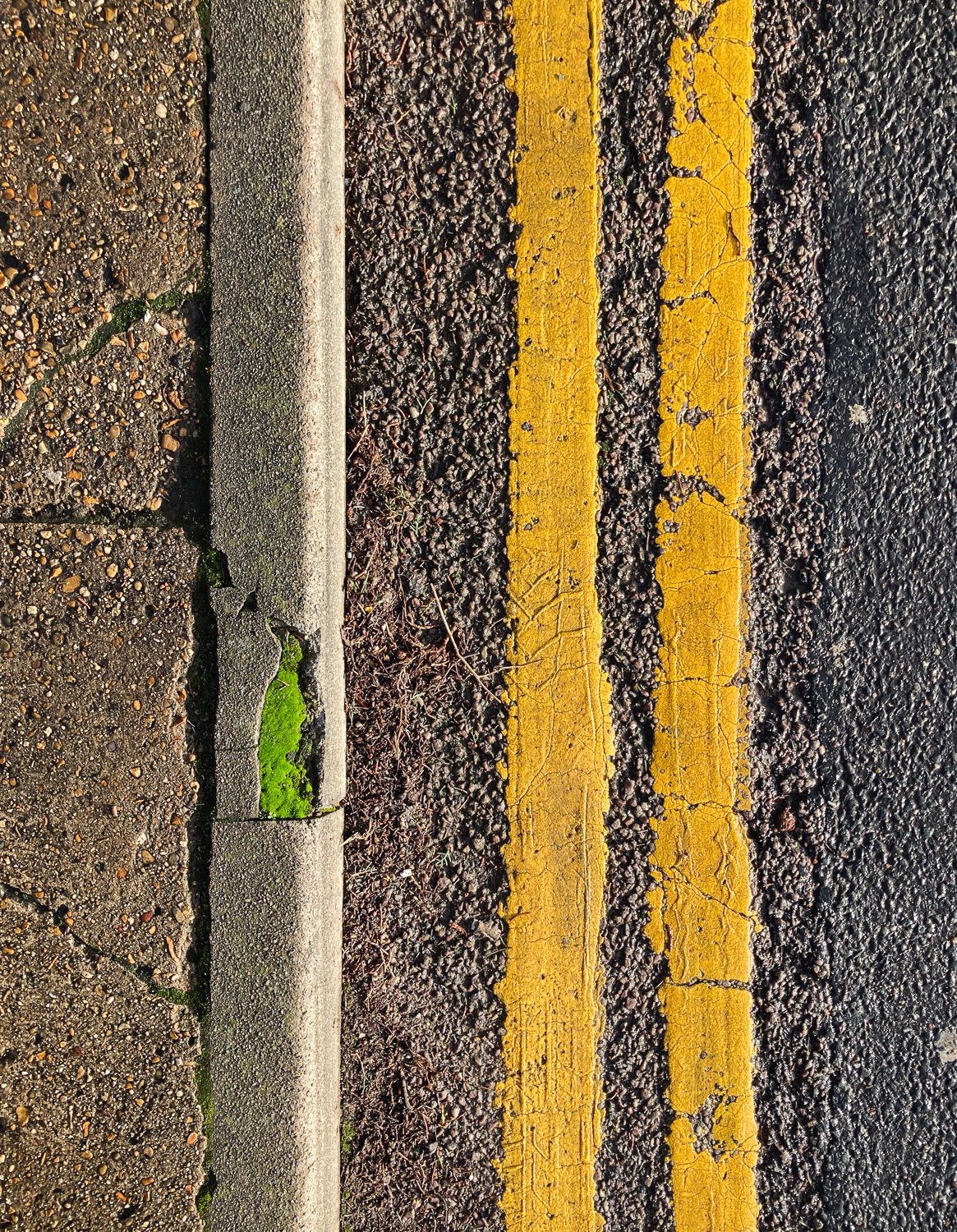 Photograph looking down on the edge of a pavement, with double yellow lines on the road to the right and the pavement and curb to the left. Part of the curb is missing and filled with bright green moss.