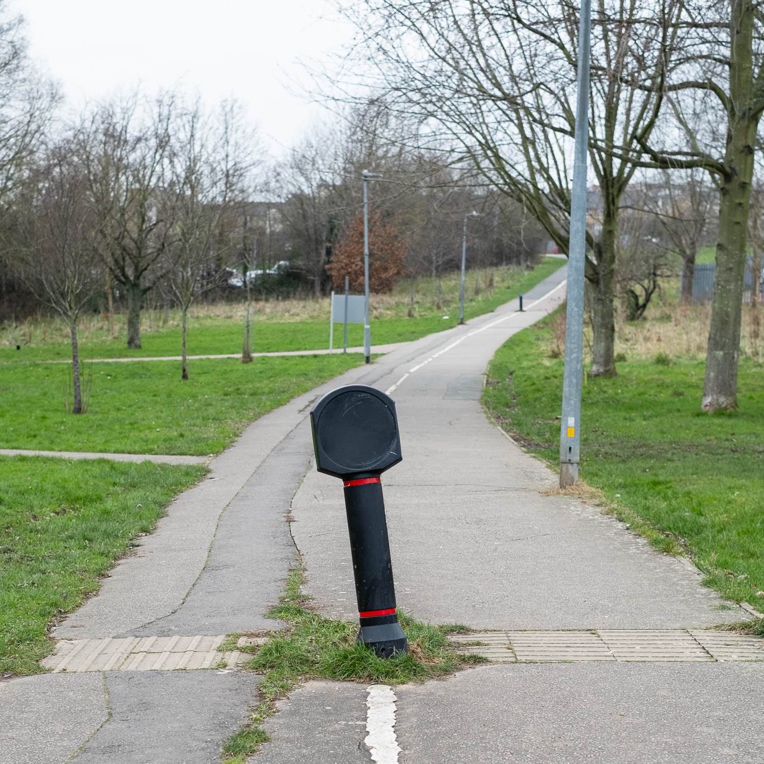 Photograph of a wide concrete path with a black bollard/sign in the centre, knocked off centre so the top leans to the left. Along the path is grass and bare trees as it is winter. The path leads off into the distance up a sligh hill.