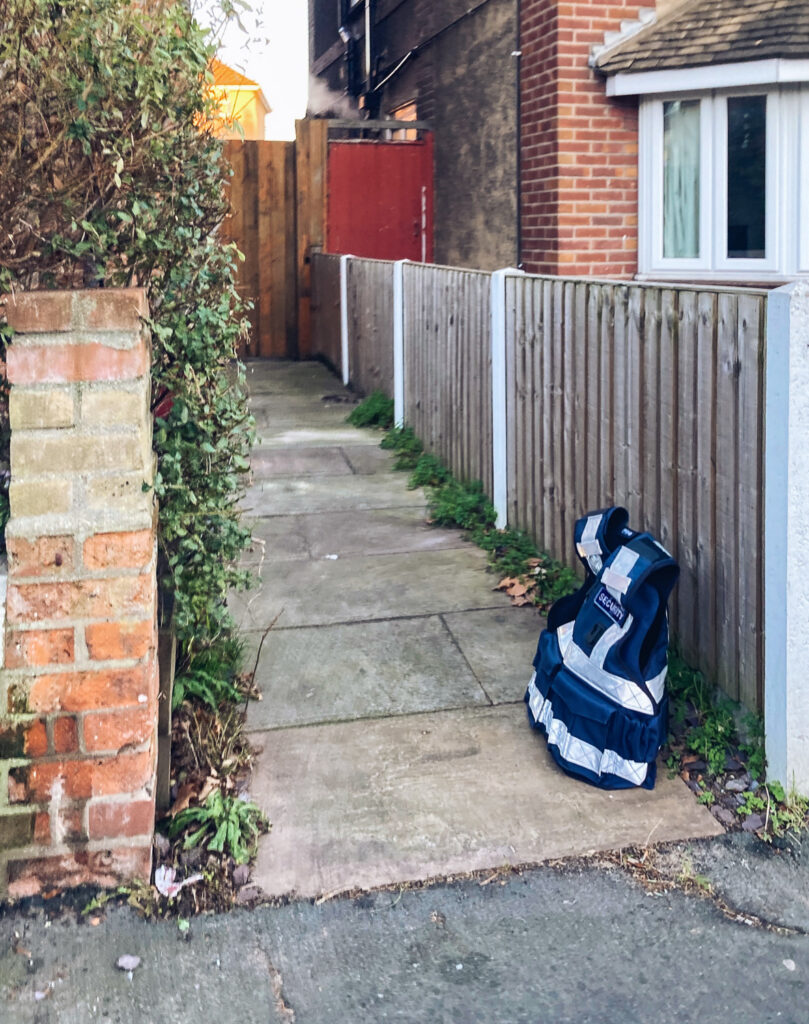 Photograph of a thick security vest in dark blue with reflective tape and a badge saying 'SECURITY', on the floor in a paved passageway between two houses, leaning on a short fence. Part of a house can be seen to the right and there's a brick wall and hedge to the left.